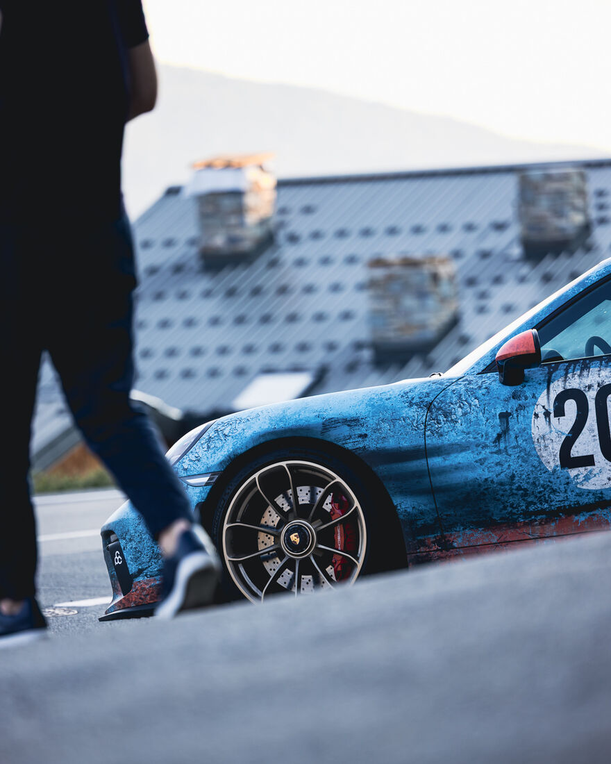 GT3 in Gulf-Lackierung, davor eine Person auf dem Parkplatz